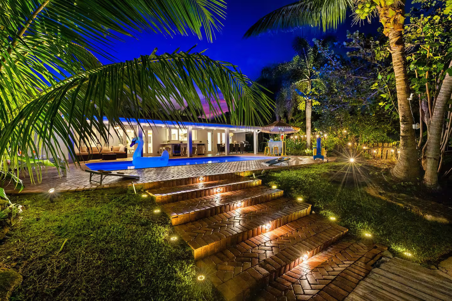 Nighttime view of heated pool, lit brick steps, pergola, and tropical garden at Pompano Beach waterfront rental